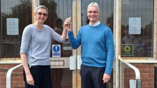 Two people standing outside, holding a set of keys between them. The woman, to the left has on a grey jumper, dark trousers and has short grey hair and glasses. The man has on a blue jumper, shirt and dark trousers. He also has glasses on and short grey hair.