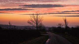 A sunset sky in shades of pink, red and orange with dark grey clouds. A skyline of trees and hills is in silhouette against the sky, and in the foreground a road winds towards the horizon.