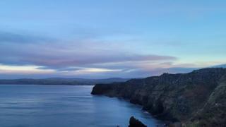 Blue sky and mauve coloured clouds sit over a blue sea inlet with a stretch of land to the right