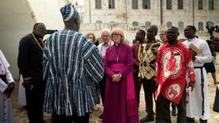 Dame Sarah Mullally, a woman with short blond hair and a fringe, wearing reddish-purple robes and a gold cross necklace. She stands in front of sand-coloured buildings and is surrounded by half a dozen Ghanaian men.