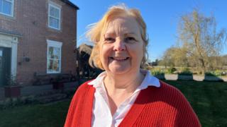 A woman with blonde hair, a white shirt and red cardigan smiles as she stands in front of a building and garden.