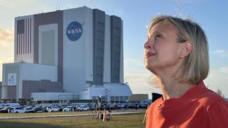 BBC Science Editor Rebecca Morelle stands about 100 metres in front of the cuboid Nasa mission control building. She is looking into the Sun as a shadow falls across the grass between her and the building. Some camera tripods are visible in the middle ground and a car park. Rebecca wears a red blouse or dress.