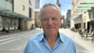 Chris wears a light blue collared shirt and has a mild smile on his face, while standing outside on a sunny day surounded by buildings.
