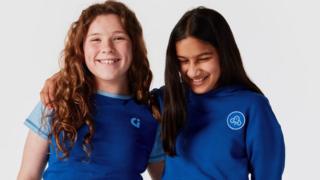 Two girls stand next to one another with their arms around the other's shoulder. The one on the left has curly ginger hair while the one on the right has dark, straight hair. They are both smiling. The girl on the left is wearing a short sleeved dark blue top with light blue cap sleeves and a small Girlguiding logo on the front. The other girl is wearing a royal blue hoodie with a circular Girlguiding logo patch on the front. The background is plain white.