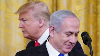 Photo showing US president Donald Trump and Israeli Prime Minister Benjamin Netanyahu. Behind them is a yellow curtain. Trump is behind Netanyahu facing left. Netanyahu is face on and looking down at a piece of paper.