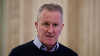 Conor Murphy, grey hair, blue shirt with dark sweater. In Stormont hall
