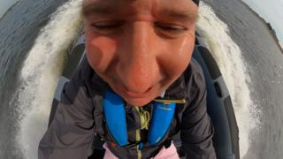 Wide angle close up of a man's smiling face, sitting in a speeding boat with wake and whitewater around the boat.