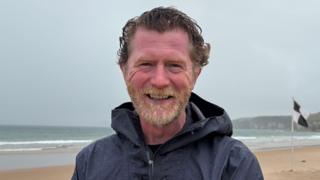Andrew Todd stands on Whiterocks beach on Portrush. He has wavy brown hair and a ginger/grey beard. He is wearing a dark blue raincoat. It is an overcast day.