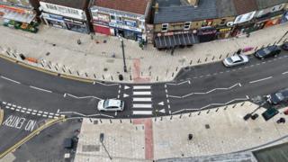Drone image of the road- with newly painted wite road markings and white pavements seen on either side of it, with cars and people can be seen using the road.