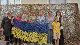 A group of women and two children standing in front of a very large tan and green camouflage net. They are holding up another small net, weaved in the blue and yellow colours of the Ukrainian flag