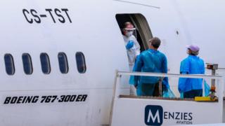 Man in full PPE talks to man and woman wearing blue plastic outer clothing and face masks at the entrance to a Menzies Aviation Boeing 767 plane.