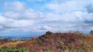 The top of a hill with fields in the far background and blue skies and clouds overhead.