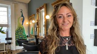 Inside a hair salon, a woman with blonde wavy hair smiles straight into the camera, her eyes sparkling. In the background is a painting of a peacock on a wall, next to big golden mirrors and black seats for customers.