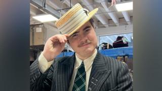 A person in a dark pinstripe suit, cream shirt, and green plaid tie stands indoors holding a straw boater hat. Behind them are shelves filled with hats and boxes in a workshop‑like space with exposed beams and fluorescent lighting.