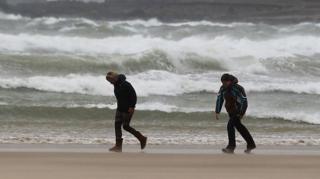 Two people bend into the wind as they walk along a beach with crashing waves behind them