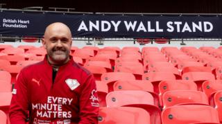 Andy Wall sitting in the Andy Wall Stand at Wembley Stadium. He is looking directly at the camera and smiling and is wearing a red zip-up long-sleeved jacket which says EVERY MINUTE MATTERS on it in white lettering. He is sitting on a red seat. Above him is a sign which says ANDY WALL STAND in white lettering.