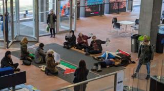 Protesters with signs and flags sit and lie on the floor of a modern building with a stone floor.