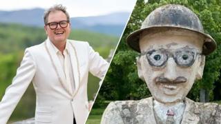 Alan Carr, wearing dark glasses and a white blazer, and a cement statue of a man in a bowler hat and round glasses