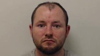 A poorly lit, grainy police mugshot of James Thomson, who has short dark hair and a beard. He is staring expressionless at the camera. Thomson is wearing a grey t-shirt and has been photographed against a grey background, without the use of a flash.