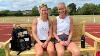 Two women sitting in running outfits on a bench with a running track behind them.