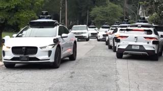 A litany of Waymo cars take up all the space in a street.