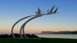 A sculpture of flying birds sits over a sea inlet with blue sky above and orange and pink sunset colours