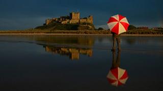 man stands with his back to camera with a big red and white umbrella open. Behind a castle sits on a hill reflecting sunshine, blue sky above and everything reflected in the shallow sea in the foreground