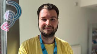 Pierce is a young man with short, brown hair and facial hair. He is smiling at the camera with his mouth closed. He is wearing a yellow t-shirt and a blue lanyard with Eastside Partnership logo on it. Behind him is a blank wall.