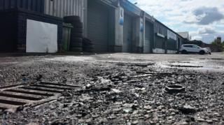 Low-angle view of an industrial estate with a row of shuttered garage units and workshops. The foreground shows a damaged asphalt surface with a drain cover, loose debris and potholes, while several parked vehicles sit beside the buildings under a partly cloudy sky.