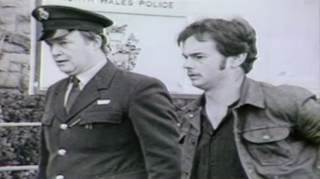 Black-and-white picture of a prisoner (on the right) being escorted by a prison officer. The prisoner is wearing a denim jacket and has a moustache.