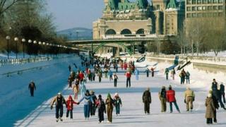 Lots of people skate along the Rideau Canal in Ottawa, Canada, which is becomes a skating rink in winter. A large castle-like building sits on the horizon.