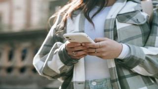 A person with long hair and with rings on their fingers holds a mobile phone in their hands. Their face is not visible.