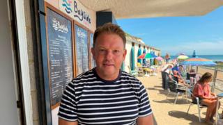 A man with short brown hair, wearing a black and white striped top by the beach.