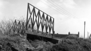 A black and white image of a small bridge with high-sided metal railings on either side and a grass hill