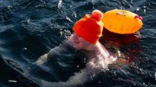 James is wearing an orange bobble hat and swimming shorts as well as swimming socks while swimming in very cold water