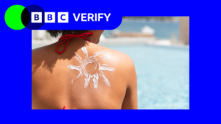 a view of a woman's shoulder from behind with sun cream lotion in the shape of a sun on her skin. she is next to a pool on a very sunny day