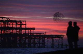 Two people watch the sky filled with red streaks as the sun rises on the winter solstice. Out into the sea is the outline of a building structure and to the right is a murmuration of birds