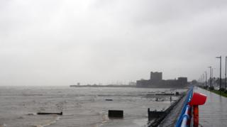 A stormy seaside promenade with rough waves, grey sky, and a distant castle in the rain.
