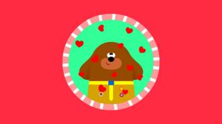 Duggee from Hey Duggee, a brown cartoon dog, in a round badge frame surrounded by hearts.
