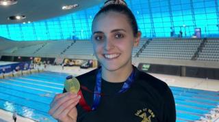 Laura Kinley in a swimming arena holing a gold medal. She is wearing a black T-shirt with the Isle of Man logo on it, and the swimming pool is behind her.