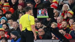 A referee in a yellow top stands in front of a football crowd while using VAR to make a decision