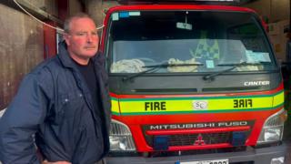 Finbar Gallagher is standing next to a red fire engine. The words fire is displayed across a green line on the vehicle. Gallagher has a navy jacket and jumper on.