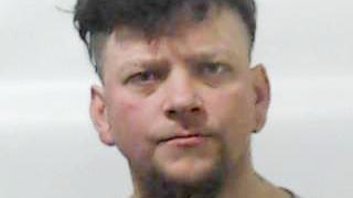 A police picture of a man with dark hair with a floppy fringe and a light goatee beard staring straight into the camera