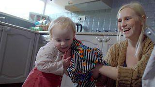 A toddler girl holding a t-shirt to her face as her mum looks on smiling.