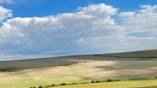 Bright blue sky with full white clouds create patches of light and shade over rolling fields below.