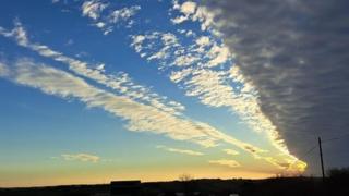 Bright blue sky over Cornwall is crossed by white diagonal streaks of golden cloud with the sun setting to the right