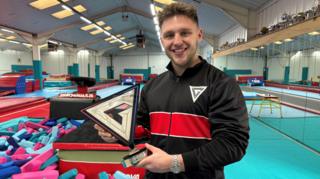 Joe Fishburn, who has dark hair and a stocky build. He is holding a triangular golden trophy in the shape of a letter G.