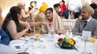People sit around a large table with a white tablecloth and centre setting of candle and red and yellow flowers in a marquee, with drinks in front of them. A woman with long black hair is talking to a woman in a head covering. A man in a suit and turban looks on. Others can be seen at tables behind, some wearing turbans and head coverings.