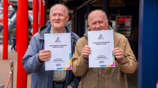 Two twin men, in their 60s, are smiling looking away from the camera. One is wearing a blue mac, the other a beige one. They are holding up white certificates close to their faces. The certificates read "20,000 rides on the big one, congratulations".
