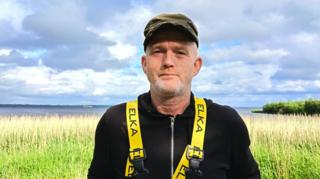 Gary McErlain is looking at the camera. He is wearing a black fleece with yellow straps over his shoulders. He is wearing a brown flat cap.